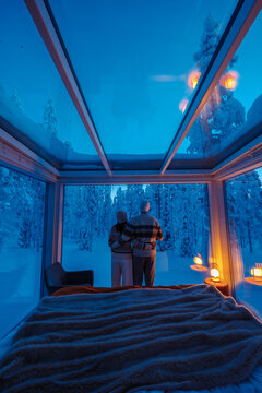 Couple enjoys a peaceful moment in a cozy cabin in Akaslompolo, Lapland. Soft blue light illuminates the snowy trees outside while they gaze through the glass walls, embracing the beauty of winter.