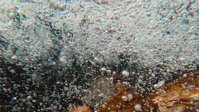 Camera dives under surface, plunging from split-level view into heart of churning mountain stream. In slow motion, chaotic storm of air bubbles becomes liquid galaxy, swirling in crystalline flow