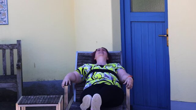 Woman enjoying sunbathing and relaxing on a wooden chair outside her home