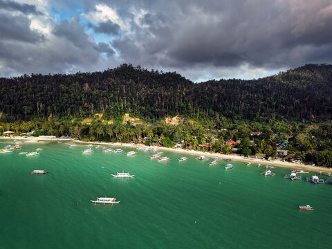 A scenic aerial view of Port Barton, Palawan, Philippines, showing a lively tropical coastline dotted with traditional banca boats anchored in calm emerald waters.
