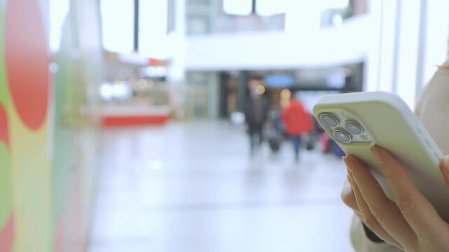 Hand holds phone in bright indoor space. Background shows blurred people walking. Light reflects off floor and walls. Focus stays on phone and hand. Scene captures modern urban environment