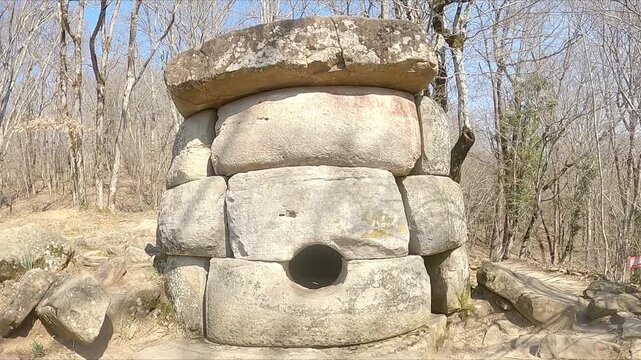 The ancient stone ruins of the dolmen stand in an open field. The weathered blocks form a disparate structure. Trees frame the background with bare branches.