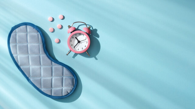 Pink alarm clock, sleep mask and scattered pink pills on a light blue surface. Concept of quality sleep routine, bedtime medication, circadian rhythm disorders and insomnia.