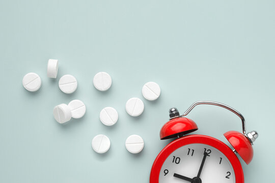 Red alarm clock with white pills on light gray background, symbolizing scheduled medication intake and health care routine. Top closeup view.