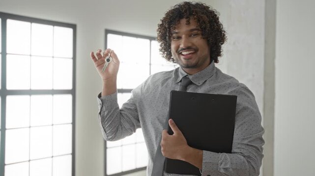 Man holds keyring in hand and clutches clipboard while smiling in building near large window and white wall; confidence trust.
