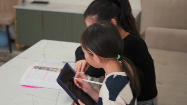 Girl completes test tasks on tablet and workbook while mother supervises, home assessment and exam preparation process, focused learning environment in Kyrgyz Asian family setting