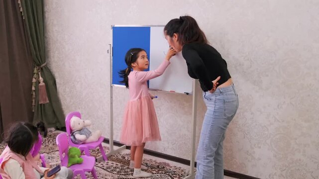 Mother teaching young Asian girl math on whiteboard at home, guiding learning process, checking answers, child education, family teaching, early school preparation and development
