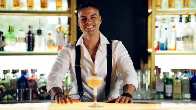 Professional bartender standing at illuminated bar counter, smiling at camera, presenting a carefully crafted cocktail