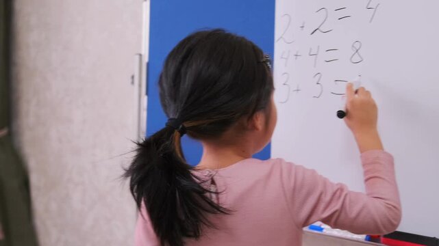 Young Asian girl solving math problems on whiteboard at home, early education concept, child learning numbers, arithmetic practice, school preparation, focused study activity indoors