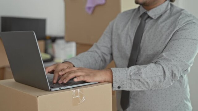 Man typing on laptop with hands on keyboard in a building amid moving boxes and unpacked belongings, wearing shirt and tie; focus.