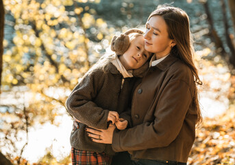 Fototapeta premium Heartwarming moment: loving mother hugging her little daughter wearing earmuffs in beautiful autumn forest.