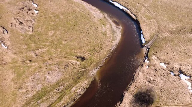 Aerial scene of a winding river bending through dry brown grasslands and melting snow patches. Top-down flyover camera angle in bright daylight. High quality 4k aerial footage.