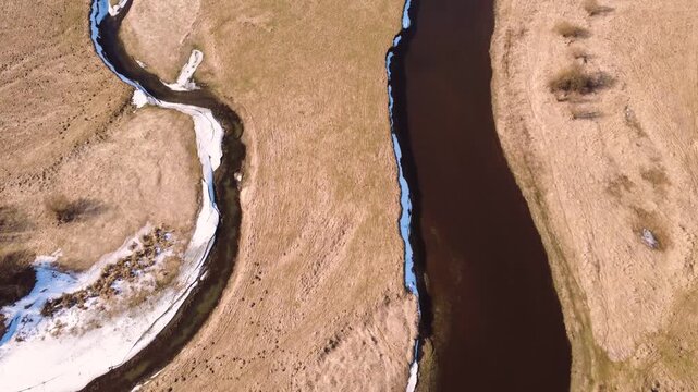 Top-down aerial tracking shot flying over a meandering river cutting through a dry brown grassy terrain with patches of melting snow under bright daylight. High quality 4k aerial footage.