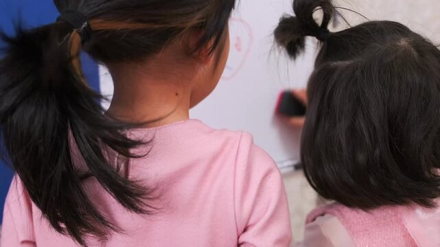 Two young girls standing at whiteboard writing and erasing, learning activity at home, early education concept, children studying together, development skills, indoor classroom play