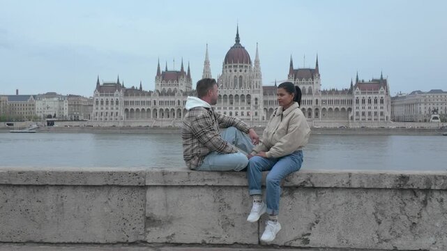 A couple shares a tender moment sitting by the Danube River. The stunning backdrop of Budapests iconic parliament building enhances their vacation experience. Perfect for travel enthusiasts.