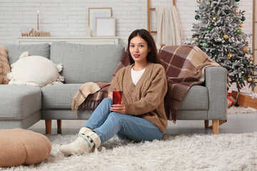 Fototapeta premium Beautiful young happy Asian woman with cup of hot mulled wine sitting on floor at home