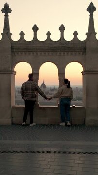 A couple enjoys a serene sunset view from Fisherman Bastion in Budapest, Hungary. They hold hands, taking in the stunning cityscape, creating a perfect moment of tranquility and connection.