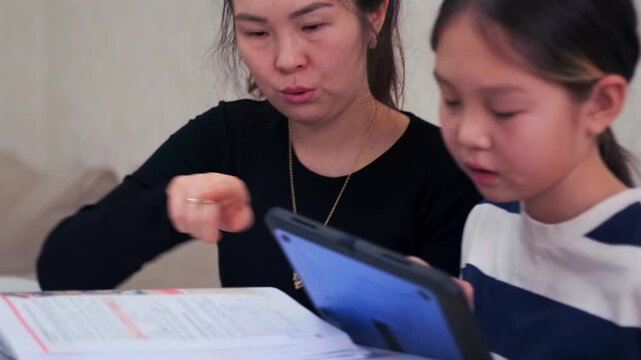 Tutor helps young girl prepare for exams using textbook and tablet, focused study and guidance, effective learning process and knowledge improvement in Kyrgyz Asian home environment