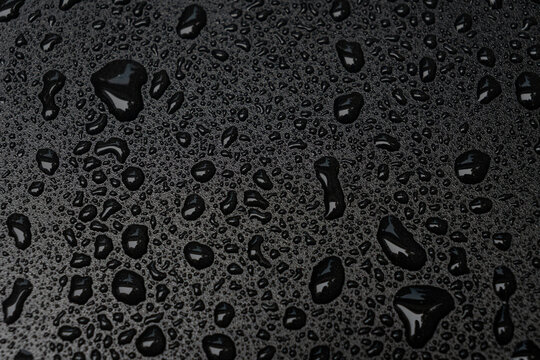 A close-up abstract view of water droplets beading on a dark waterproof surface with glossy reflections and strong texture.