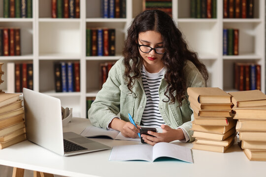 Female student in eyeglasses using mobile phone and writing in notebook at table with laptop and stack of books in library