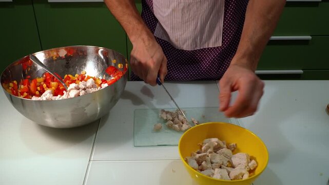 Male hands dicing cooked chicken on cutting board in kitchen, adding pieces to bowl with vegetables. Close-up of food preparation process, homemade meal cooking, protein ingredient and healthy diet