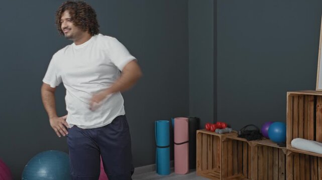 Man stretching side with hand on hip and extended arm in studio gym, surrounded by mats, exercise balls and dumbbells; serenity focus.