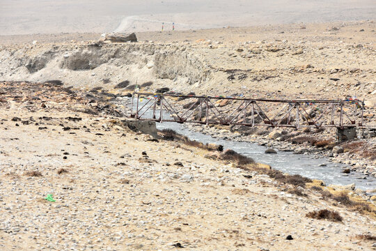 Rustic metal bridge spanning a narrow mountain stream in a barren high-altitude landscape, with rocky terrain and remote dirt tracks under a pale sky in North Sikkim.