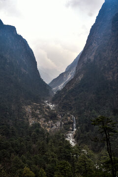 Scenic Himalayan valley with cascading waterfall flowing through dense pine forest, surrounded by steep mountains and rugged rocky terrain in North Sikkim.