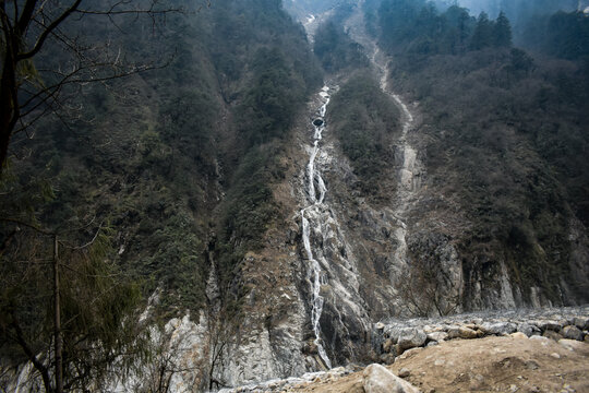 Tall cascading waterfall flowing down a rugged forested mountainside, surrounded by mist, rocky cliffs, and dense Himalayan greenery in North Sikkim. 