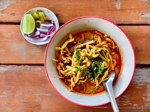 Traditional Northern Thai curry noodle soup (Khao Soy) with tender beef, topped with crispy noodles. Served with lime, red onions, and pickled cabbage on a wooden table.