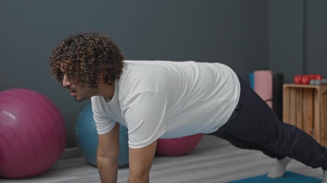 Man on blue yoga mat in studio doing plank with shoulder tap beside pink exercise ball and wooden crate; focus core strength balance.