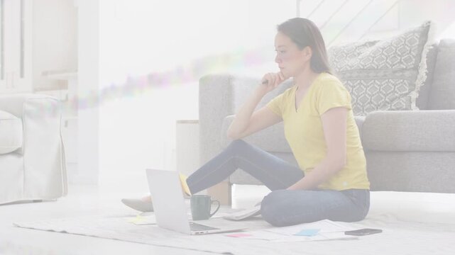 Mid adult woman starting study session on rug, reviewing laptop and papers, examining handheld item