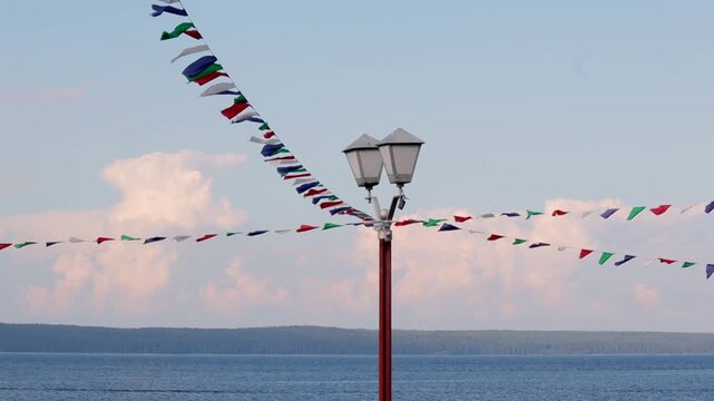 String of multicolored festive flags stretches across the frame, dancing in the breeze against the serene expanse of lake. Classic street lamp stands in the foreground, adding a touch of charm to the 