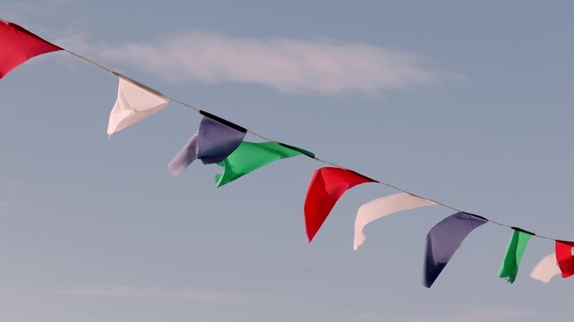String of triangular flags in vibrant hues - red, white, green, and blue - dances in the breeze against a serene blue sky. The dynamic movement of the flags creates a cheerful, celebratory atmosphere,