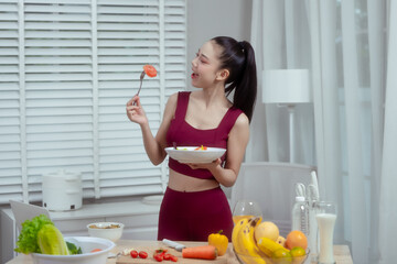 Obraz premium Young woman in burgundy activewear eats salad at kitchen counter with fresh fruit and vegetables, smiling in bright modern home, healthy lifestyle and diet concept.