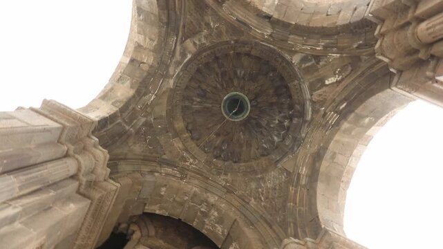 A bottom up view looking into the vaulted stone ceiling and arches of an ancient historic structure. The intricate masonry features a central hanging bell against bright sky.