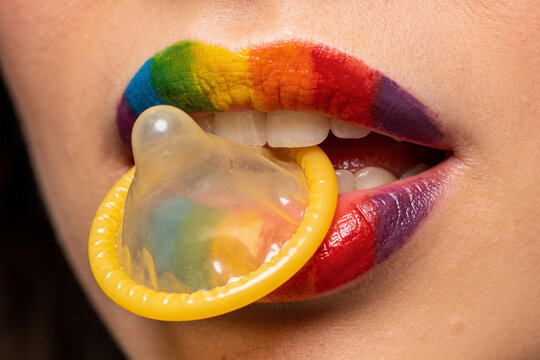 Close-up. A woman holds a condom in her mouth, her lips are painted in rainbow colors.