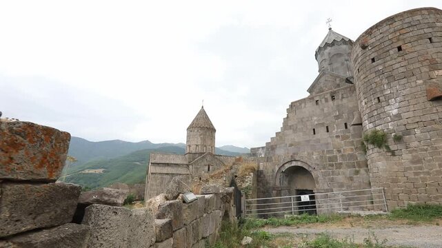 Scenic view of the historic Tatev Monastery nestled in lush green Armenian mountains, transitioning to a close-up of ancient carved cross-stones resting against the majestic stone building walls.