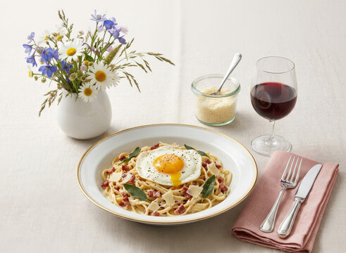 Linguine &agrave; la parisienne with a fried egg, crispy bacon, sage, and a glass of red wine, served on an elegant table with flowers