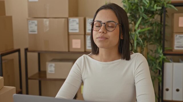 Woman with parcels and cardboard boxes smiles at camera while seated at a packing desk in building; small business confidence.