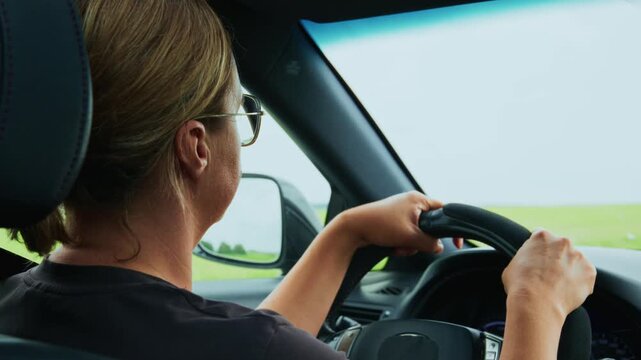 Over-the-shoulder view of woman driving car on open road. She wears glasses, focused, hands on wheel. Scene shows calm, safe, travel or commuting theme. Good for travel, road safety