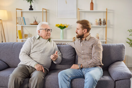 Serious senior father sitting on sofa and having meaningful conversation with his adult son. Two men sharing thoughts and engaging in discussion during friendly family dialogue in living room at home.
