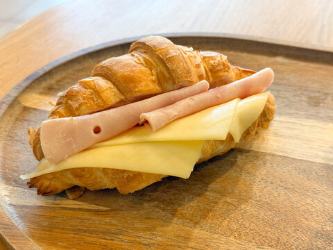 Close view of croissant sandwich with cheese and ham on wooden tray. Breakfast choice, simple meal, baked product texture, and food preparation detail in cafe setting.