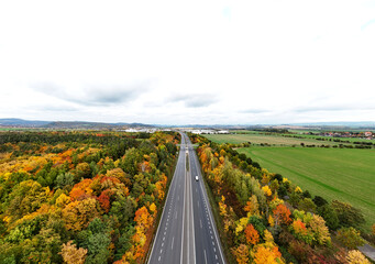 Naklejka premium Wide aerial view of a straight highway passing through a vibrant autumn forest and green agricultural fields under a cloudy sky. Concept of travel and infrastructure.