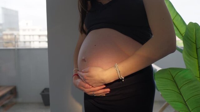 Expectant woman cradles round belly with both hands, bracelet and ring visible on wrist, green plant blurred beside bright window in background