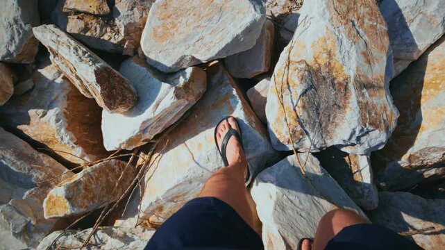 Person in flip-flops steps carefully across sunlit rocky terrain. View from above, focusing on feet navigating uneven stones. Shadows stretch across surface, indicating bright daylight
