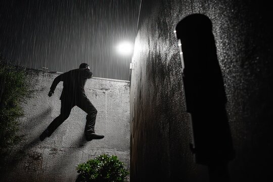Man leaping over wet concrete wall in heavy rain at night under a streetlight, shadowed vertical post and small bush nearby, tense and stealthy mood