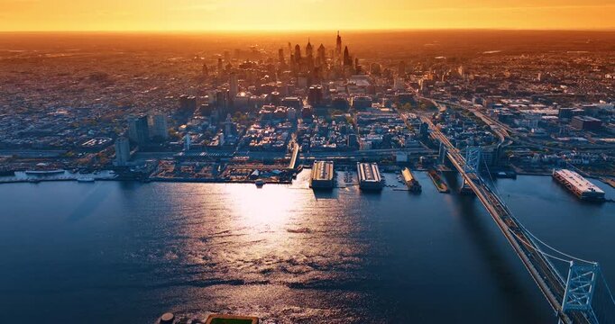 Warm golden light of sunset covers the scenery of Philadelphia, Pennsylvania, USA. Drone footage over the waterfront of the Delaware River near the Benjamin Franklin Bridge.