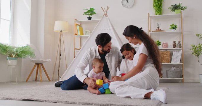 Happy family playing with block toys together, sitting on carpet in cozy living room with teepee. Carefree parents and children play learning game, teamwork of father and mother, baby toddler and girl