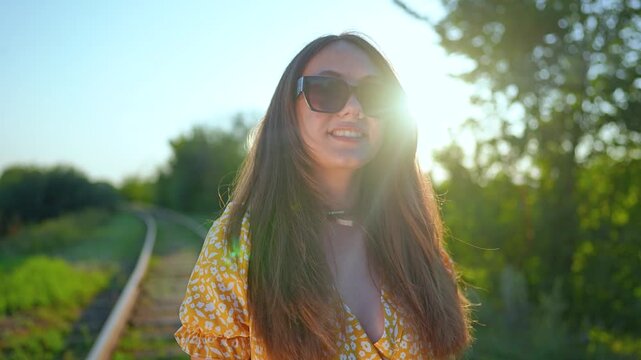 Happy young woman stands near railway track at sunset. Girl in sunglasses  and yellow dress smiles outdoors. Female traveler enjoys sunny summer  evening. Beautiful woman walks near railroad.